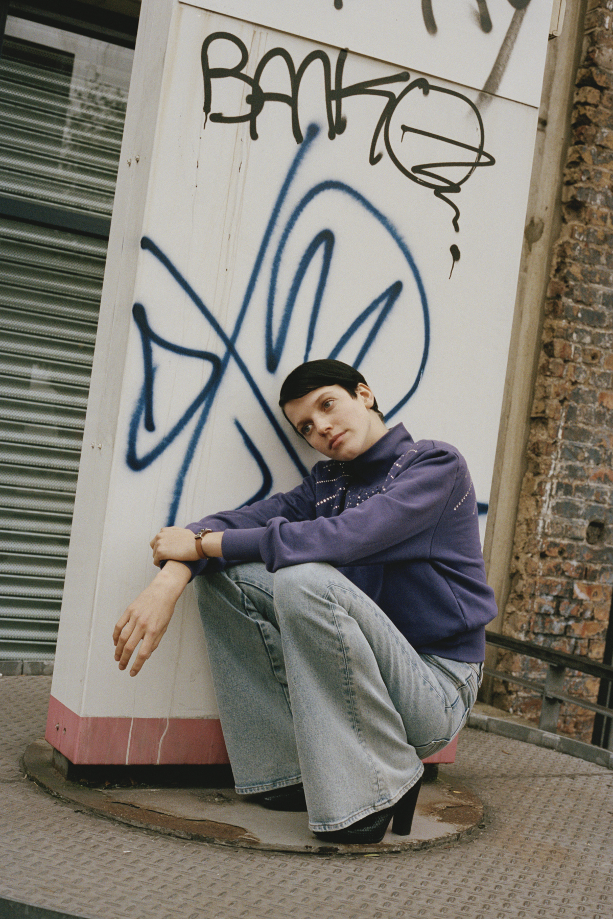 Model Yuki van Gog squatting in front of a graffiti in a purple jumper